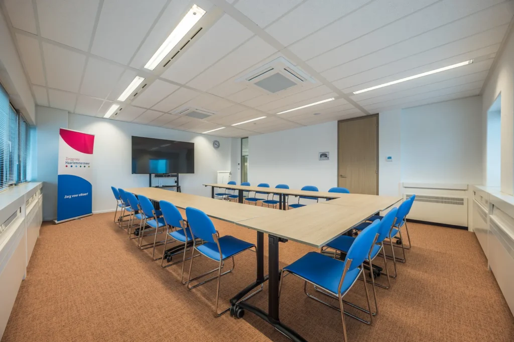 Vergaderruimte van Zorggroep Haarlemmermeer met blauwe stoelen, een grote scherm en een banner met de tekst "Zorg voor elkaar".