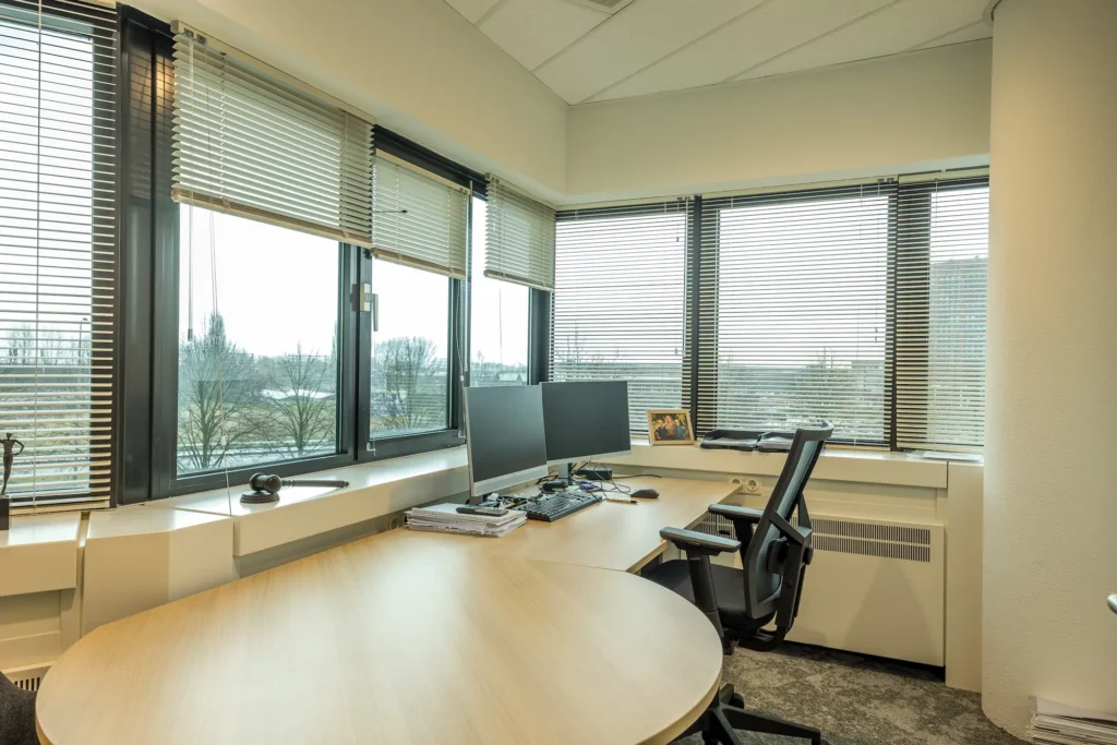 Een modern kantoor van de Zorggroep Haarlemmermeer met hoekbureau, dubbele monitoren en uitzicht op bomen.