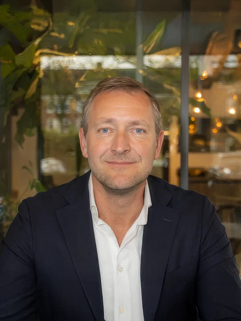 Portret van een man in een zakelijke omgeving, gekleed in een donkerblauw pak en witte overhemd, zittend voor een groene, moderne achtergrond.