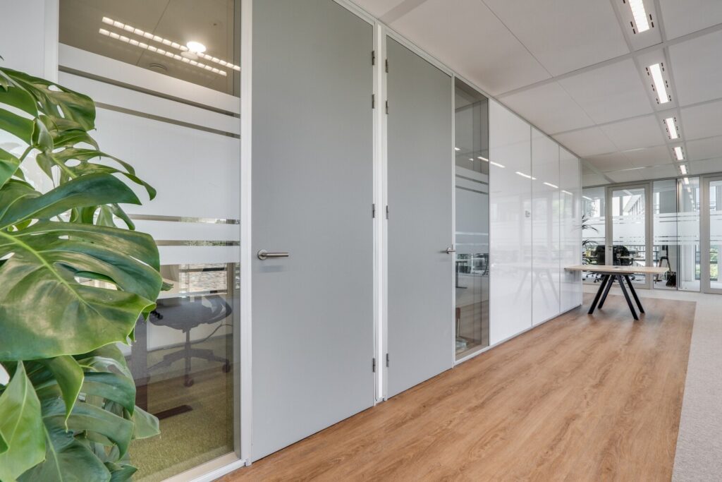 Moderne kantoorruimte met glazen wanden, houten vloer en een grote plant links.