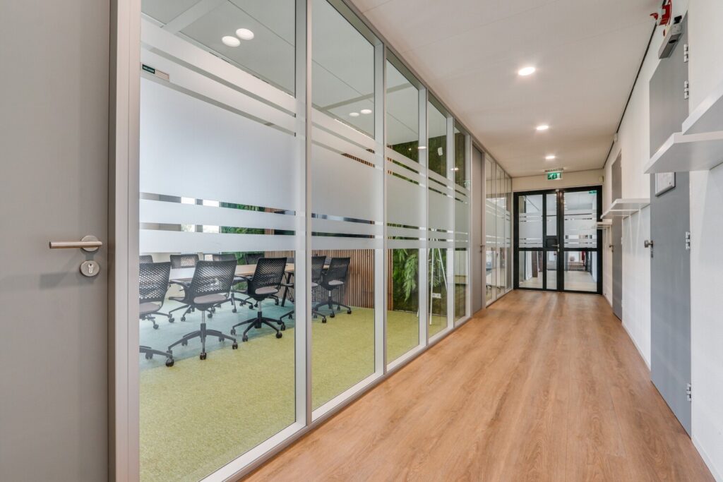 Lichte, moderne gang met glazen wanden en uitzicht op een vergaderruimte met stoelen.