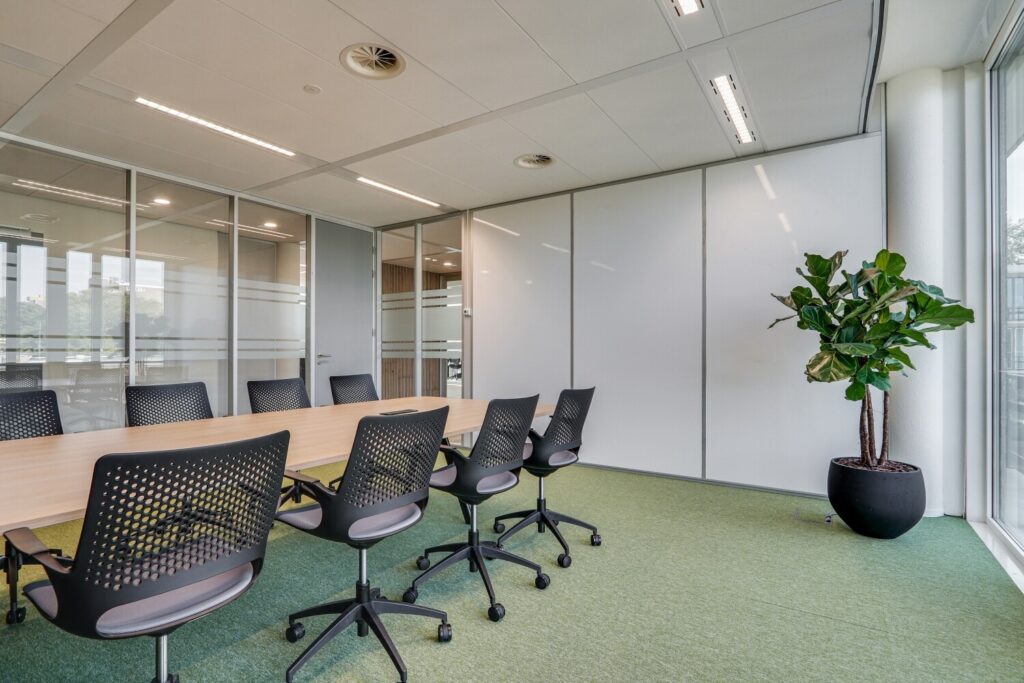 Moderne vergaderruimte met glazen wanden, een lange tafel, stoelen op wieltjes en een grote groene plant in de hoek.