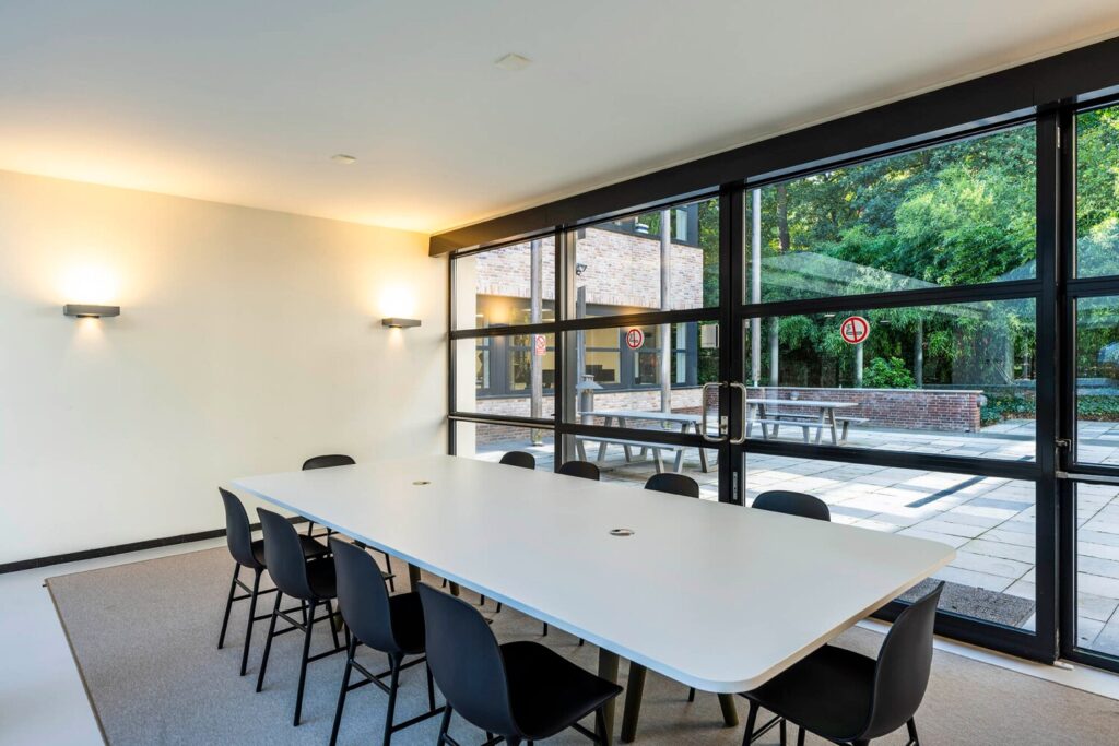 Vergaderruimte met een lange witte tafel en moderne stoelen, met uitzicht op een buitenplaats met groen en bakstenen muren.