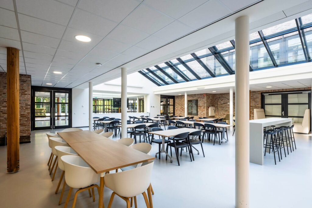 Een moderne, lichte kantine met houten en witte meubels, omringd door glazen wanden en een glazen dak.