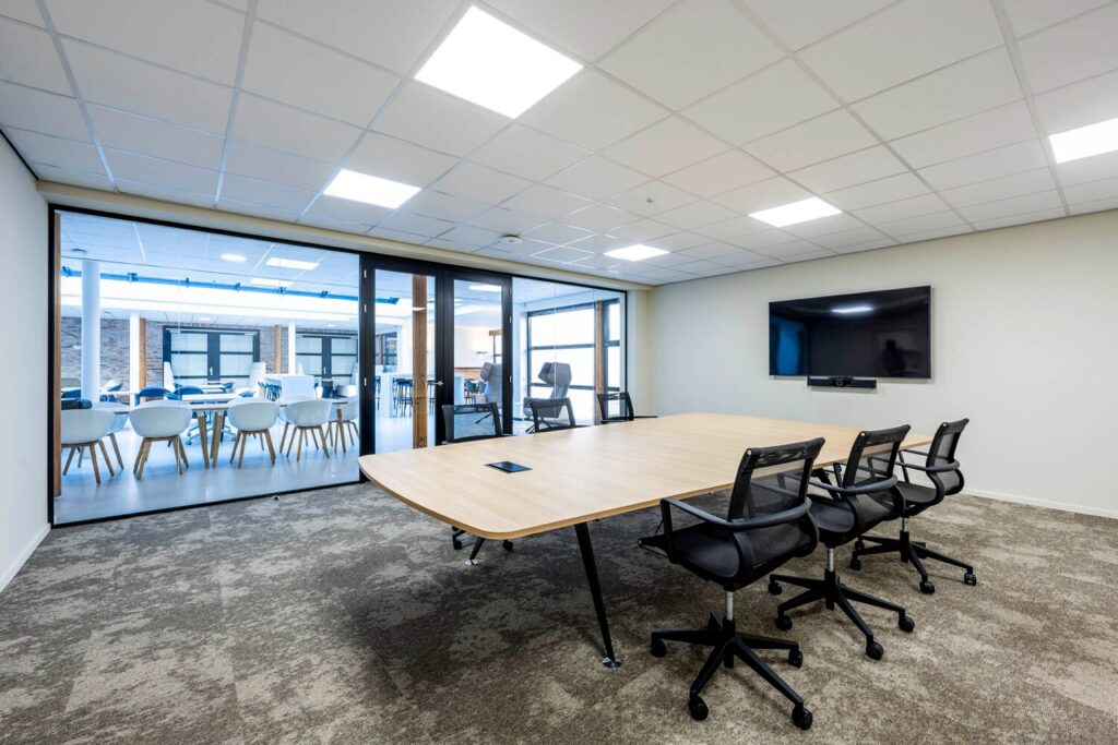 Moderne vergaderruimte met een lange houten tafel, vergaderstoelen, een groot scherm en uitzicht op een open kantoorruimte.