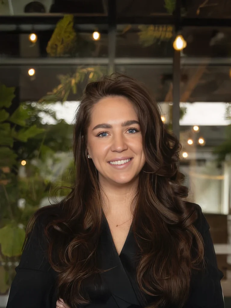 Portret van een lachende vrouw met lang, donker haar voor een sfeervolle achtergrond met groene planten en verlichting.