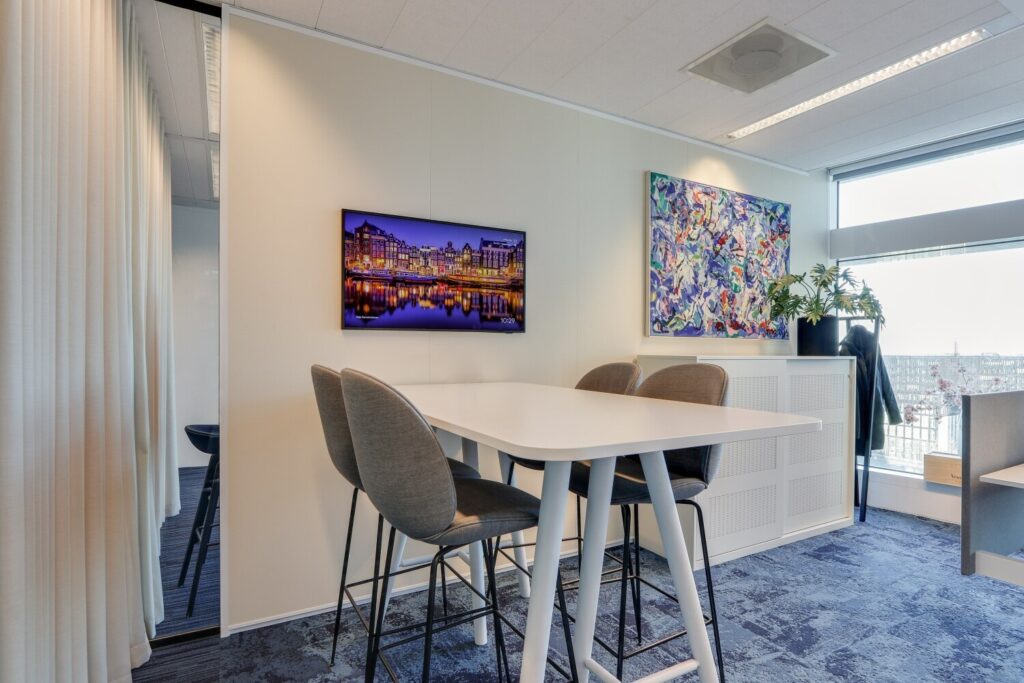 Moderne kantoorruimte met een vergadertafel, hoge stoelen, kleurrijke kunst aan de muur, en een groot raam met uitzicht naar buiten.