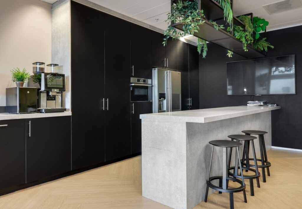 Moderne kantoorkeuken met zwarte kasten, een koffiezetapparaat, en een natuurlijke plantendecoratie, naast een grijs keukenblok met krukken.