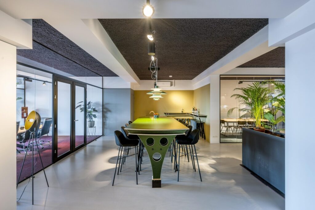 Een moderne kantoorruimte met glazen wanden, een lange tafel met hoge stoelen en veel planten.