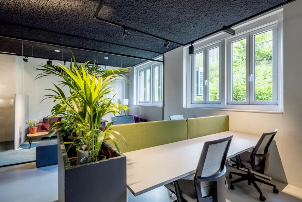 Modern kantoorinterieur met bureaus, comfortabele stoelen en veel groen, met grote ramen die natuurlijk licht binnenlaten.