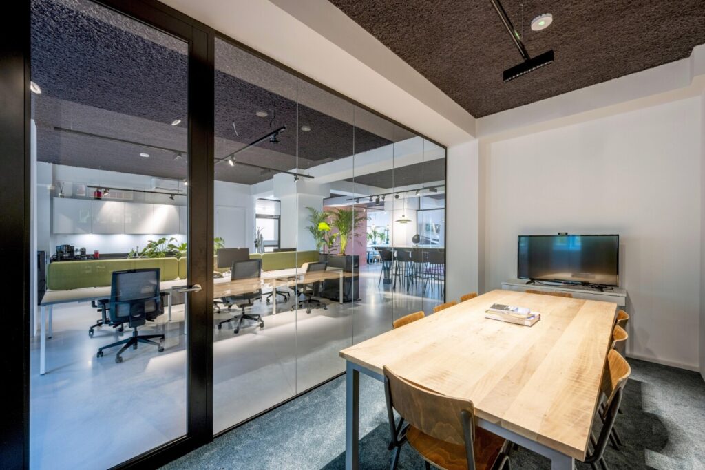 Moderne vergaderruimte met een houten tafel en zes stoelen, naast een glazen wand die zicht biedt op een open kantoorruimte.