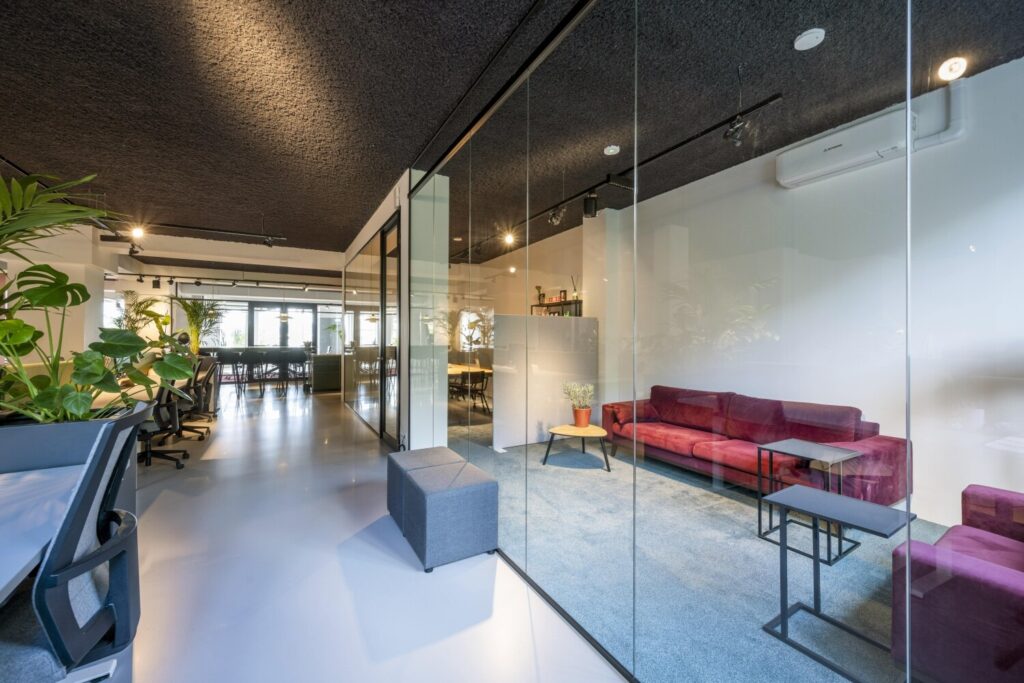 Een moderne kantoorruimte met glazen wanden, ruime werkplekken, een rode bank en veel groenplanten.