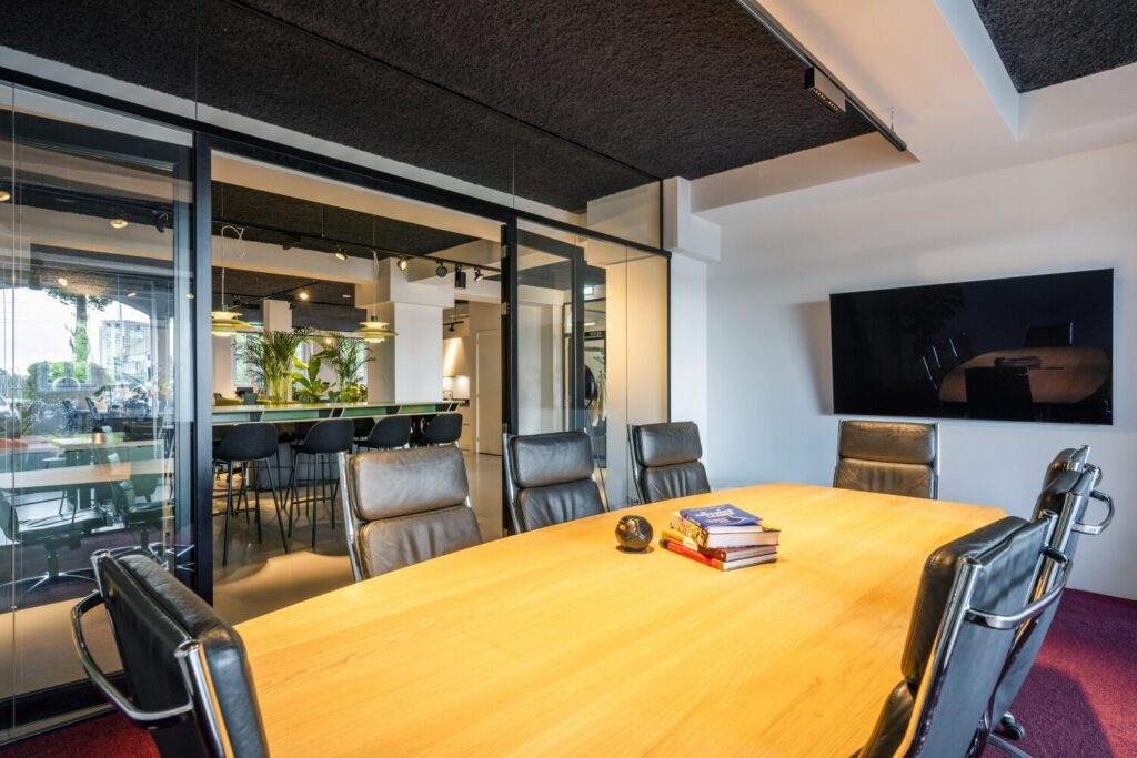 Moderne vergaderruimte met een grote houten tafel, lederen stoelen, en een televisie aan de muur, naast een stijlvolle kantoorruimte.