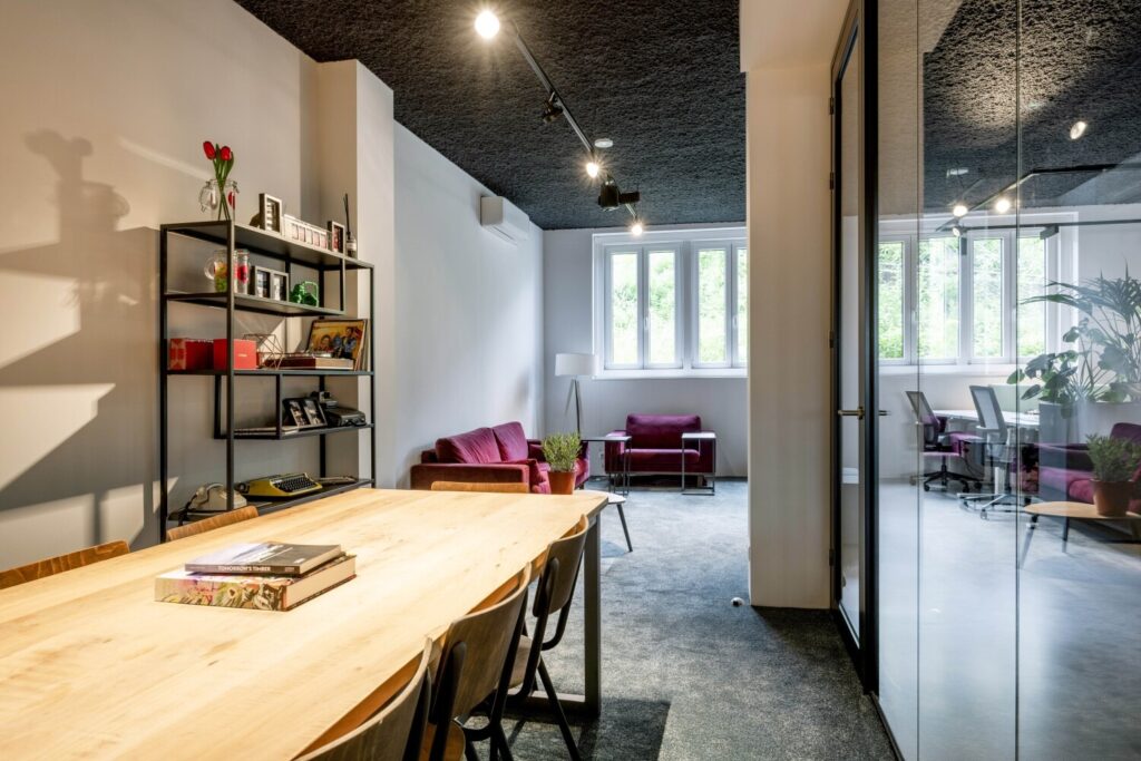 Stijlvolle moderne kantoorruimte met houten tafel, kleurrijke planken en een comfortabele zithoek bij een groot raam.