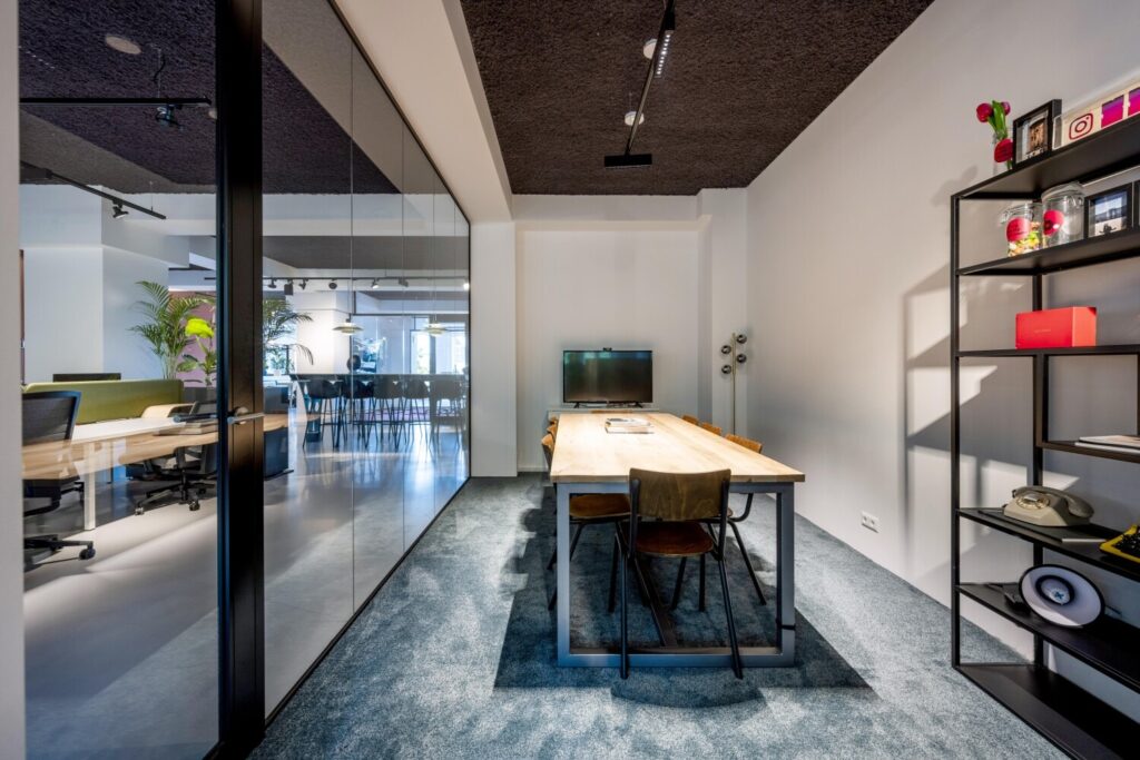 Moderne vergaderruimte met een houten tafel, stoelen, een televisie en een open kast, grenzend aan een kantoor met glazen wanden.