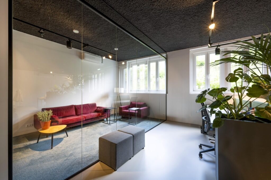 Moderne kantoorruimte met glazen wand, voorzien van een rode bank en planten, en veel natuurlijk lichtinval.