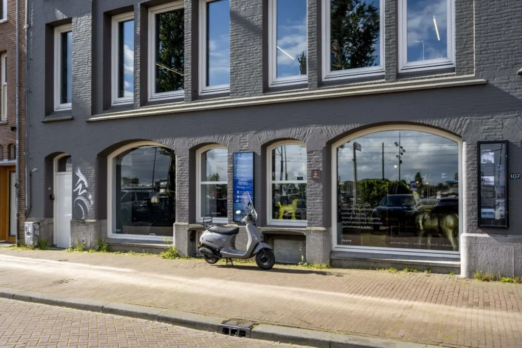 Grijs bakstenen kantoorgebouw met grote ramen en een scooter geparkeerd voor de ingang.