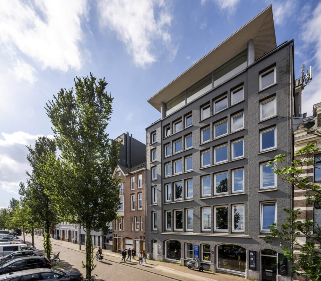 Modern kantoorgebouw met grote ramen langs een boomrijke straat in een stedelijke omgeving.