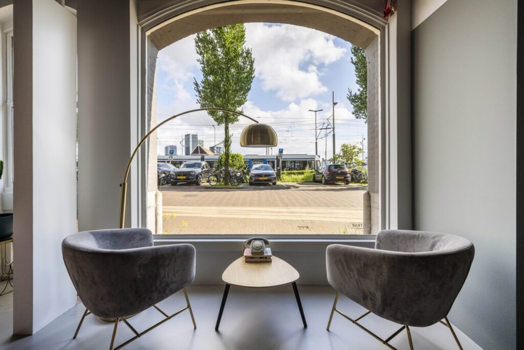 Twee grijze fauteuils met een moderne vloerlamp en een houten bijzettafel voor een groot raam met uitzicht op een straat en geparkeerde auto's.