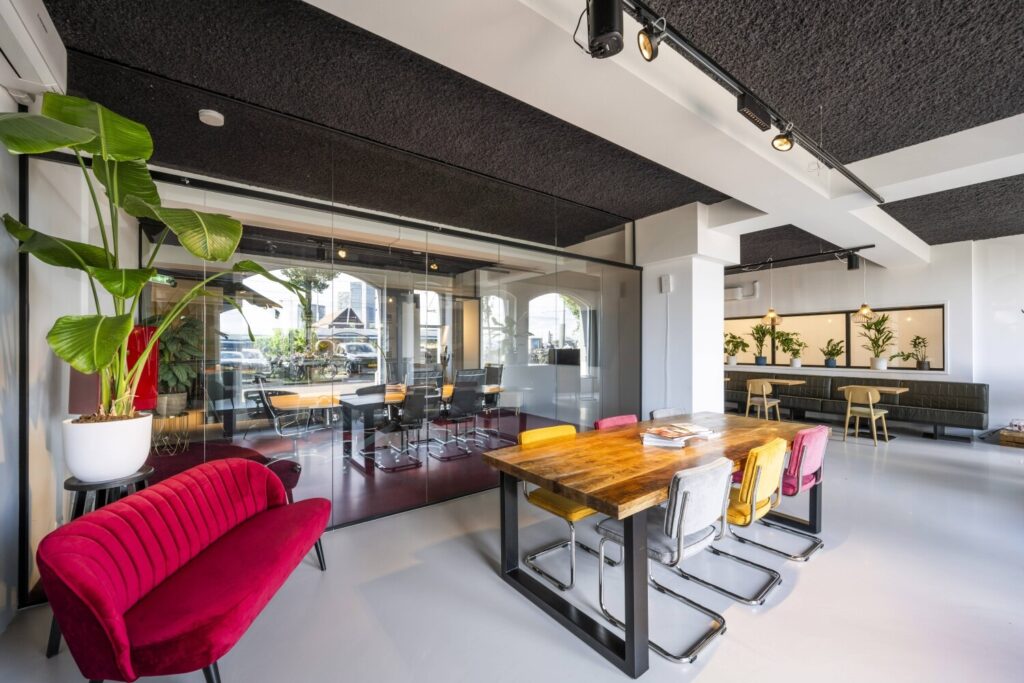 Modern ingerichte kantoorruimte met glazen vergaderruimte, kleurrijke stoelen, een houten tafel en planten.