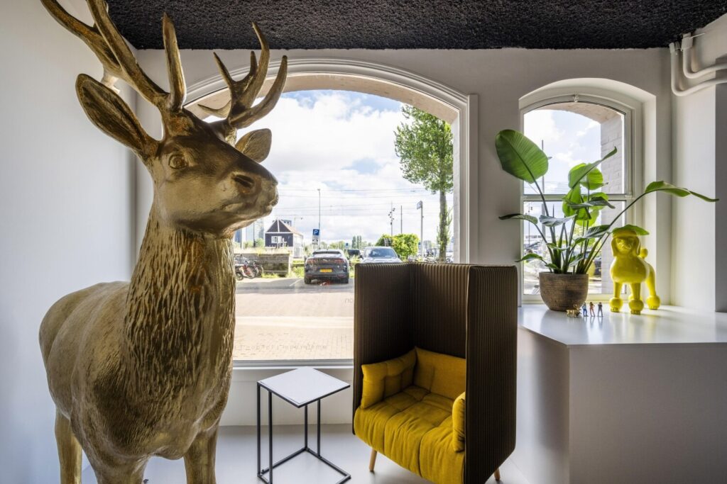 Een moderne ruimte met een groot hertbeeld, een gele stoel, en groenplant naast speels geel decor, met uitzicht op een parkeerplaats door grote ramen.