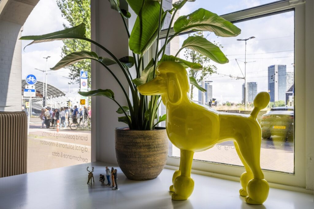Een helder gele plastic poedelstandbeeld naast een groene plant op een vensterbank met uitzicht op een drukke straat en enkele hoge gebouwen.