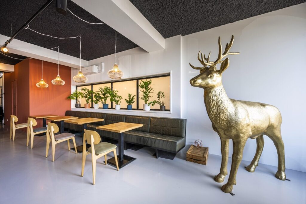 Een moderne ruimte met houten tafels, stoelen, groene planten en een groot gouden hertensculptuur.