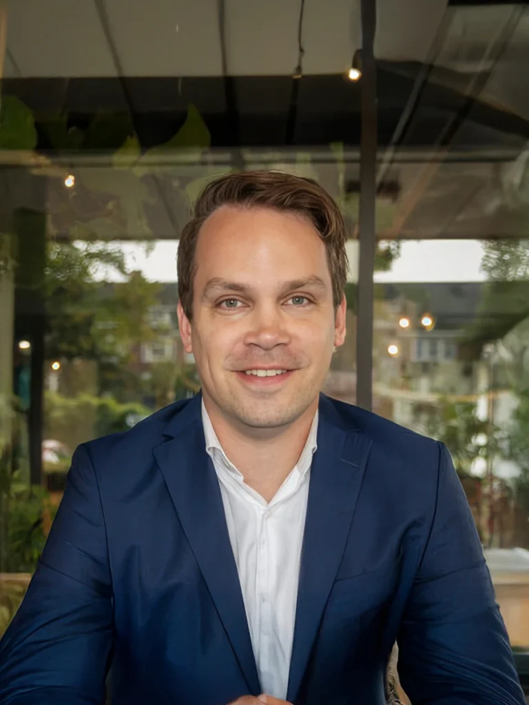 Ik kan de persoon in de afbeelding niet identificeren, maar het toont een man in een blauw pak met een witte overhemd, zittend voor een raam.