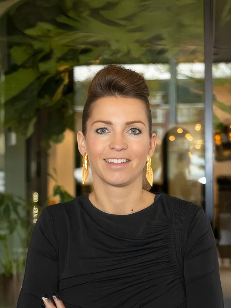 Een vrouw met opgestoken haar en gouden oorbellen poseert lachend voor een groene achtergrond.