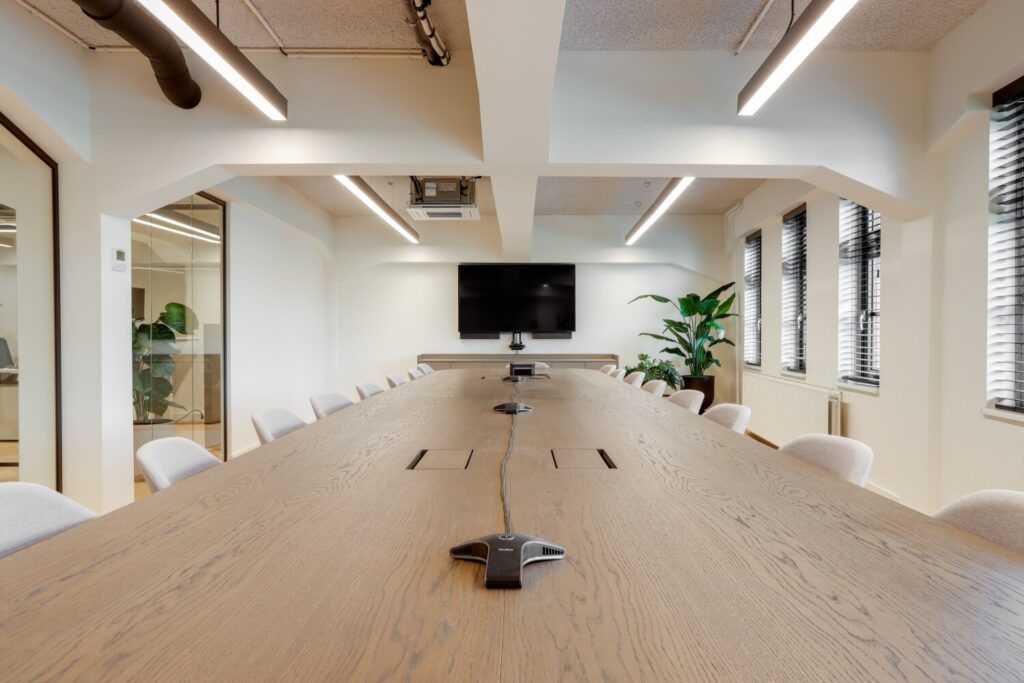 Een moderne vergaderruimte met lange houten tafel, gestoffeerde stoelen, een groot scherm aan de muur en enkele planten voor een frisse uitstraling.