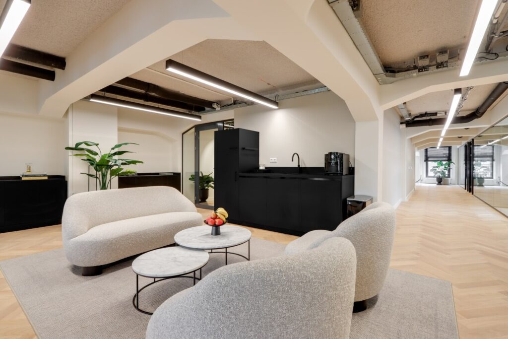Moderne kantoorruimte met zitgedeelte, ronde marmeren tafeltjes, zwarte keukenhoek en planten.