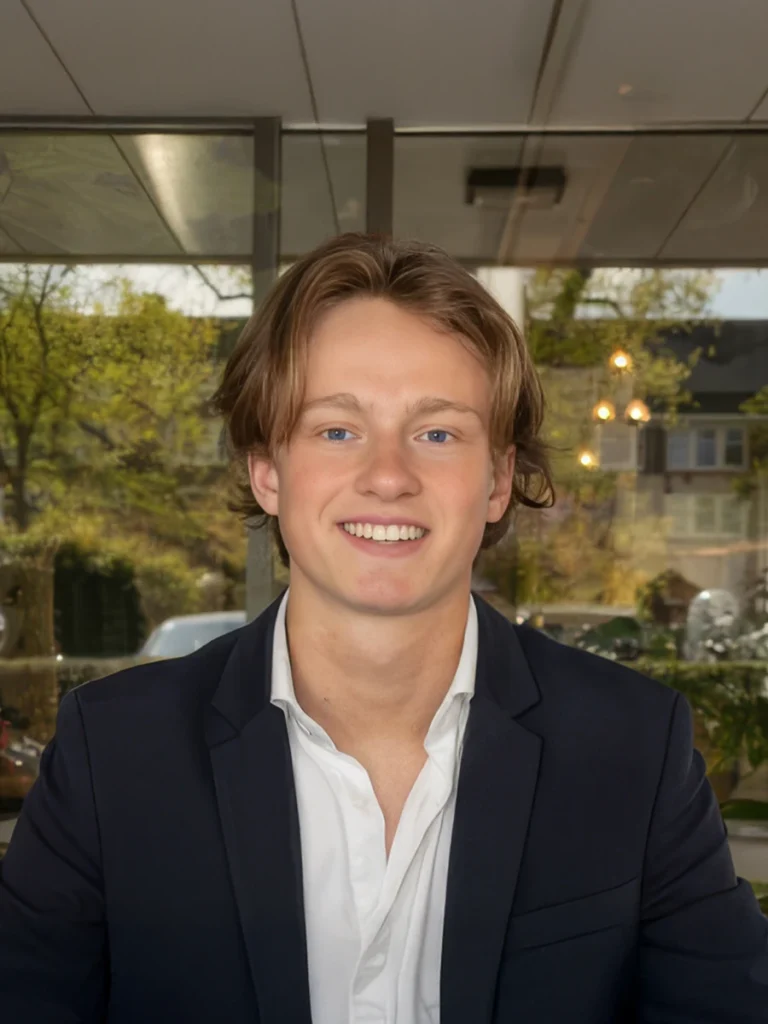 Een jongeman in een donkerblauw pak en wit overhemd glimlacht naar de camera in een moderne kantooromgeving.