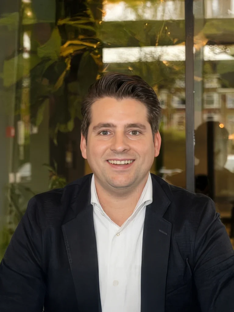 Een glimlachende man in een donkere blazer zit in een moderne kantoorruimte met planten op de achtergrond.