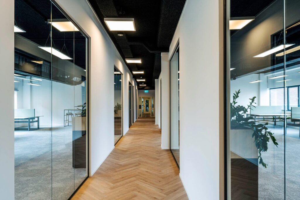 Een moderne gang met glazen wanden en houten vloer in een kantoorpand aan de Transformatorweg 76.