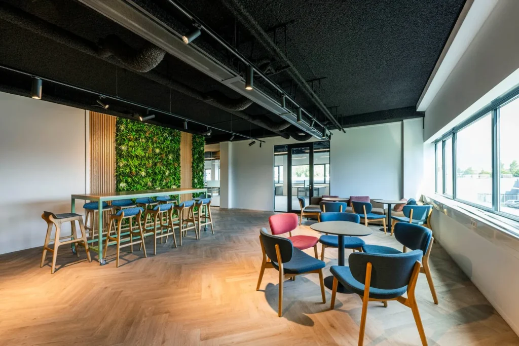 Moderne kantine bij Transformatorweg 76 met kleurrijke stoelen, grote ramen en een verticale plantenmuur.