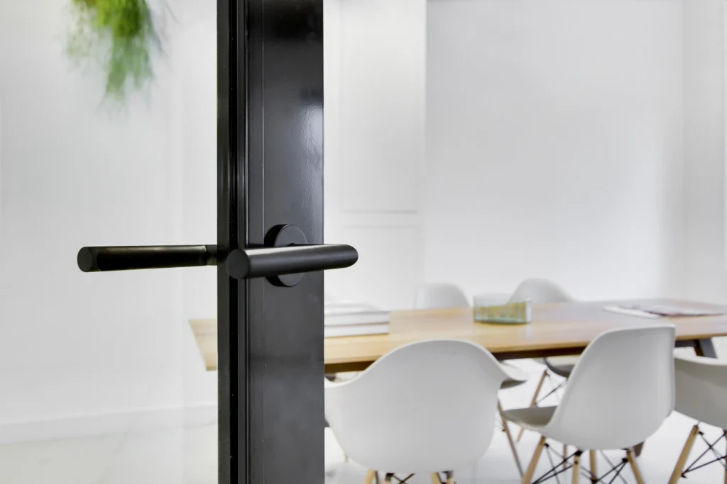 Een moderne glazen deur met zwart handvat leidt naar een stijlvolle vergaderruimte met witte stoelen en een houten tafel.