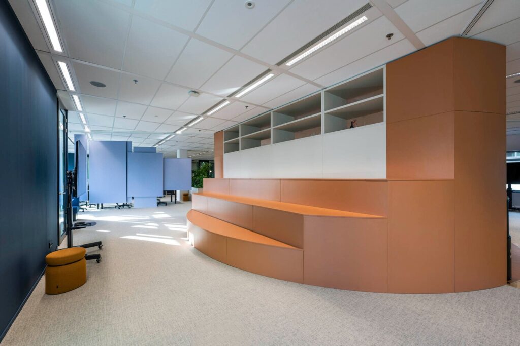 Een moderne kantoorruimte met een ronde oranje zitbank en blauwe scheidingswanden bij Winthontlaan 11.