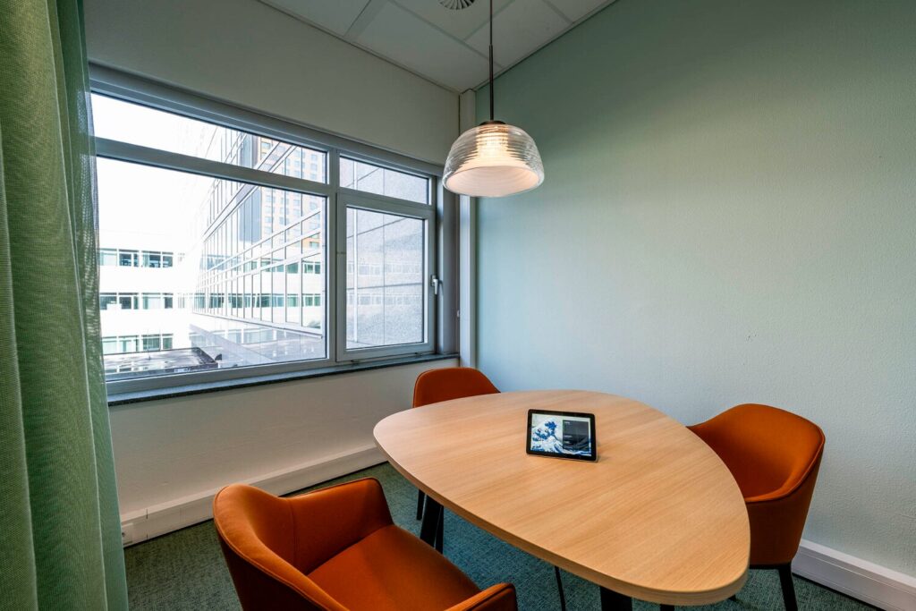 Moderne vergaderruimte met een houten tafel, twee oranje stoelen en een groot raam met uitzicht op een kantoorgebouw.